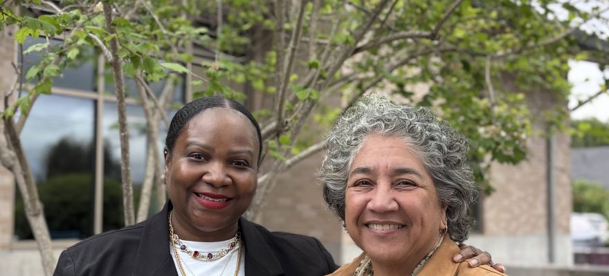 Two smiling women stand next to each other in front of a short tree with green leaves.